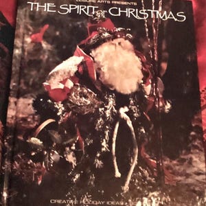 May include: A book cover featuring a red and white Santa Claus figure with a white beard, holding a sack and standing in a snowy forest. The book title is "The Spirit of Christmas" and the subtitle is "Leisure Arts Presents Creative Holiday Ideas".