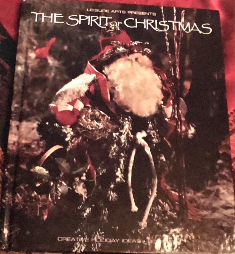 May include: A book cover featuring a red and white Santa Claus figure with a white beard, holding a sack and standing in a snowy forest. The book title is "The Spirit of Christmas" and the subtitle is "Leisure Arts Presents Creative Holiday Ideas".