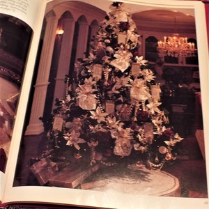 May include: A large, decorated Christmas tree with white and gold ornaments, red berries, and white poinsettias. The tree is in a large room with white columns and a chandelier.