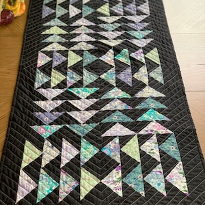 May include: A black table runner featuring a geometric pattern of triangles in shades of purple, green, and blue. The triangles are crafted from fabric with floral prints.