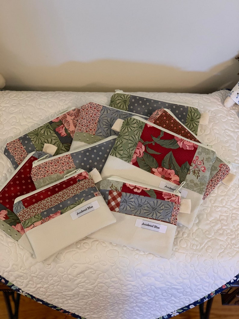 Puede incluir: Una pila de ocho peque&ntilde;as bolsas con cremallera hechas de tela de patchwork en tonos de rojo, verde, azul y blanco. Las bolsas tienen un forro blanco y un tirador de cremallera blanco. Las bolsas est&aacute;n etiquetadas con "Accidental Bliss".