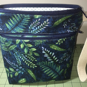 May include: A dark blue zippered pouch with a leafy pattern and a white canvas strap. The pouch has two zippered compartments.