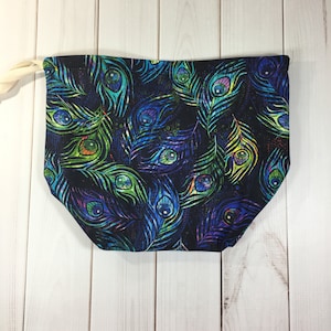 Op de afbeelding: Een zwarte stoffen trekkoordtas met een pauwenverenprint. De veren zijn in tinten blauw, groen en paars.