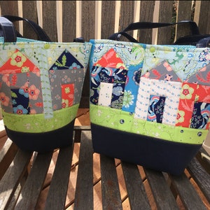 Könnte beinhalten: Zwei Tragetaschen mit blauen Henkeln und einem dunkelblauen Boden. Die Taschen sind aus Patchwork-Stoff mit einem Muster von Häusern in den Farben Blau, Grün, Rot und Grau.