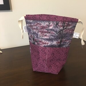Op de afbeelding: Een paarse trekkoordtas met een bloemenpatroon op de onderste helft en een boslandschap op de bovenste helft. De tas heeft een wit trekkoord.