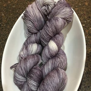 May include: Three skeins of purple and white yarn in a white bowl. The yarn is a gradient of purple and white.