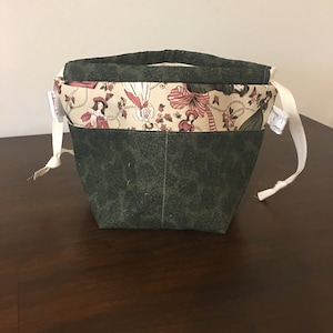 Op de afbeelding: Een groene stoffen trekkoordtas met een witte stoffen voering en een wit trekkoord. De voering heeft een patroon van vrouwen in jurken en bloemen.