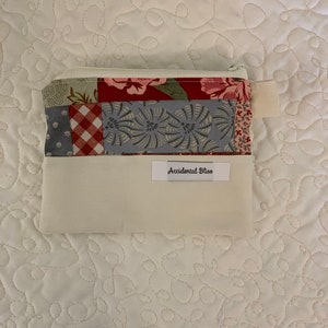 Puede incluir: Una bolsa blanca con cremallera con un dise&ntilde;o de patchwork en tonos rojos, azules y verdes. La bolsa tiene una etiqueta blanca con el texto "Accidental Bliss" impreso en ella.