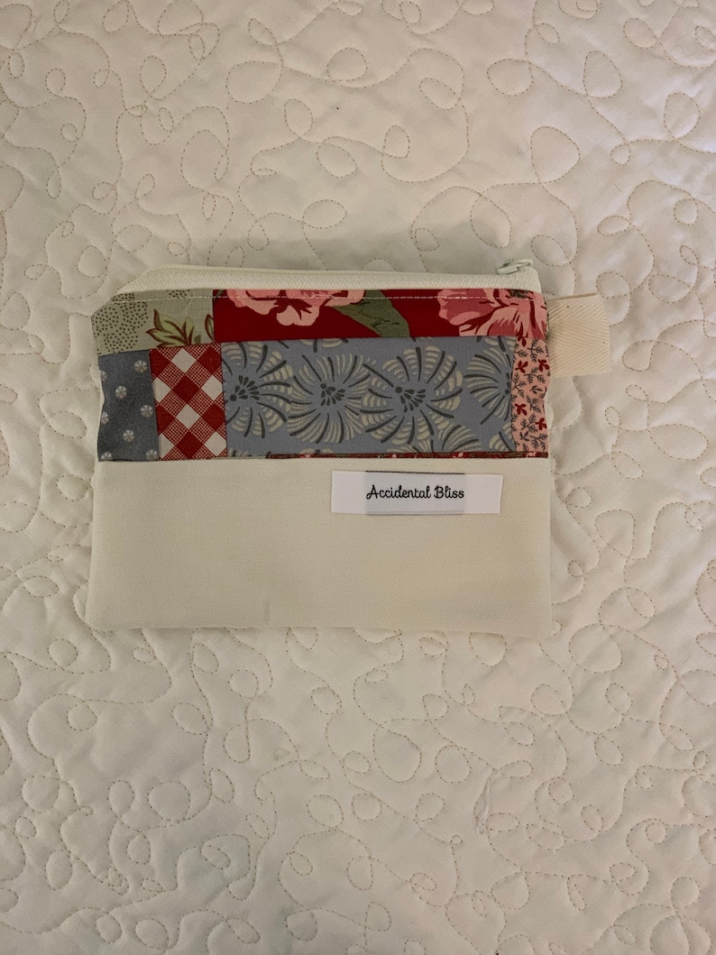 Puede incluir: Una bolsa blanca con cremallera con un dise&ntilde;o de patchwork en tonos rojos, azules y verdes. La bolsa tiene una etiqueta blanca con el texto "Accidental Bliss" impreso en ella.