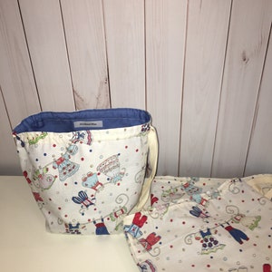 Op de afbeelding: Twee witte stoffen trekzakken met een blauwe en rode print van kinderkleding die aan een waslijn hangen. De zakken hebben een blauw label met de tekst "Accidental Bliss".