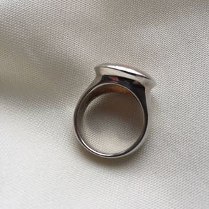 Puede incluir: Un anillo de plata con una parte superior lisa de forma ovalada. El anillo est&aacute; sobre un fondo de tela blanca.
