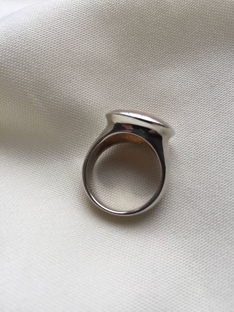 Puede incluir: Un anillo de plata con una parte superior lisa de forma ovalada. El anillo est&aacute; sobre un fondo de tela blanca.