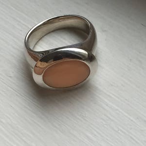 Puede incluir: Un anillo de plata con una piedra ovalada de color melocot&oacute;n engastada en el centro.