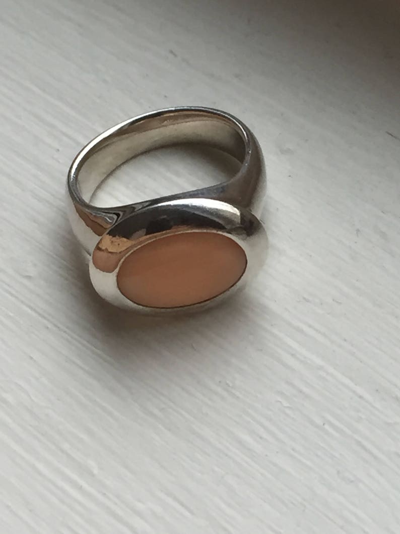 Puede incluir: Un anillo de plata con una piedra ovalada de color melocot&oacute;n engastada en el centro.