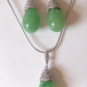 May include: A jewelry set featuring a necklace and matching earrings. The set includes a silver chain necklace with a teardrop pendant and a pair of teardrop earrings. Each piece has a light green, polished stone and is accented with a silver-colored, ornate top.