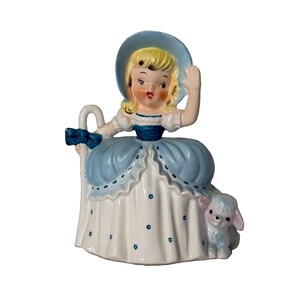 May include: A ceramic figurine of a blonde character wearing a blue bonnet and a white dress with blue floral accents and a blue waist sash. The character holds a white shepherd's crook with a blue bow and stands next to a small white lamb.