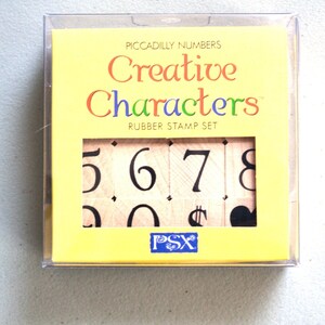May include: A yellow box with a clear plastic cover containing a set of rubber stamps. The box has the text "Piccadilly Numbers Creative Characters Rubber Stamp Set" printed on it. The stamps include the numbers 5, 6, 7, 8, 0, a dollar sign, and a heart.