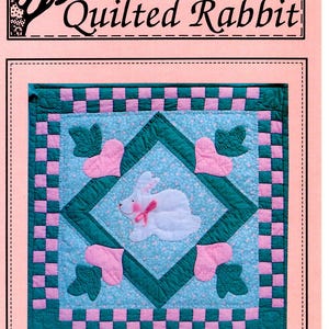 May include: A pink and green quilt pattern with a white bunny in the center. The pattern is called "The Carrot Patch" and the number is #ST-204. The quilt pattern is from The Quilted Rabbit.