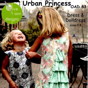 May include: Two girls wearing dresses, one in a white and black floral print and the other in a green and pink patterned dress with a ruffled bottom. The dress pattern includes a green, pink, and white floral design. The girls are standing on a brick path. The image includes the text "Urban Princess OAD 83 Dress & Dolldress sizes 2-8".
