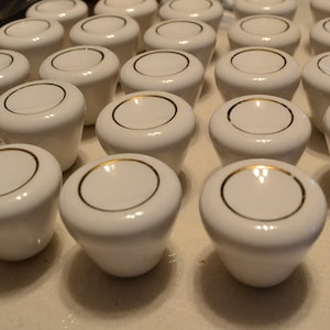 May include: A collection of white ceramic knobs with a gold trim. The knobs are arranged in rows on a white surface.