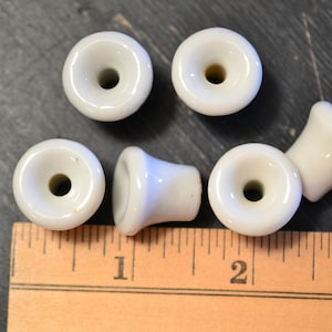 May include: Six white ceramic drawer pulls with a flared design and a central hole. The pulls are arranged on a dark surface next to a wooden ruler, with measurements in inches.