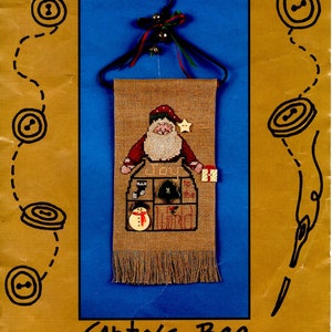 May include: A cross stitch pattern for a Santa's bag with ceramic buttons. The pattern includes a hanger, fabric, and a chart. The bag is decorated with a snowman, a stocking, a bell, and a tree. The text "To the World" is stitched on the bag.