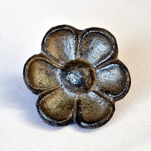 May include: A black cast iron flower-shaped knob with a raised center. The knob has six petals and a smooth, slightly shiny finish.