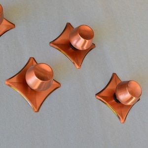 Peut inclure: Quatre boutons de cabinet en cuivre avec une base en forme de diamant. Chaque bouton a un dessus arrondi et une finition lisse et brillante.