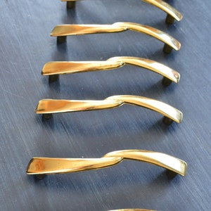 May include: A set of ten gold-toned cabinet pulls with a twisted design. The pulls are arranged in a row on a dark blue surface.