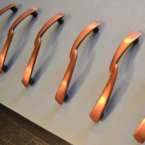 May include: Seven copper cabinet pulls with a curved, twisted design. The pulls are arranged in a row on a light blue surface.