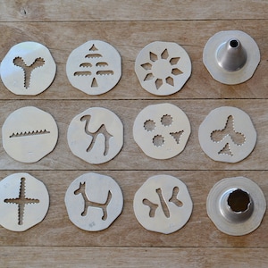 May include: A set of 12 metal cookie cutters with various designs, including a tree, a camel, a sun, a flower, and a dog. The cutters are arranged in three rows of four on a wooden surface.