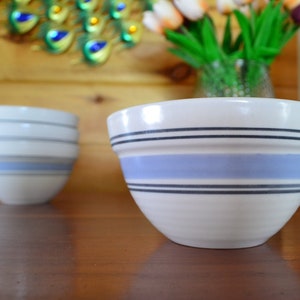 May include: Two white ceramic bowls with blue and black stripes. The larger bowl is in focus and the smaller bowl is stacked on top of two other bowls.