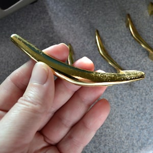 May include: A set of five gold-toned cabinet door pulls with a curved, modern design. The pulls are made of metal and have a smooth, polished finish.