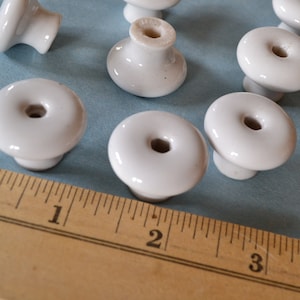 May include: A collection of ten white ceramic knobs with a raised center hole. The knobs are approximately 1 inch in diameter and are arranged on a blue surface with a wooden ruler.
