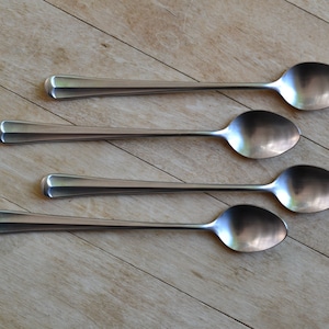 May include: Four silver spoons with long handles are arranged in a row on a light brown wooden surface.