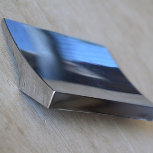 May include: A shiny, silver, rectangular cabinet pull with a curved edge. The pull is made of metal and has a smooth, polished finish.