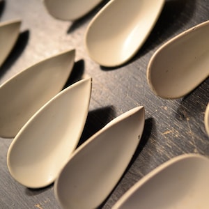 May include: A collection of ten white ceramic spoons, shaped like teardrops, arranged in a row on a dark wooden surface.