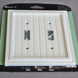 May include: White wall plate with two switch openings and a woodgrain design. The packaging says "Elumina" and "Composite Wood White".