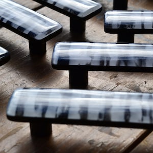 May include: A set of six black and white glass cabinet knobs with a rectangular shape. The knobs have a black metal base and a clear glass top with a white pattern.