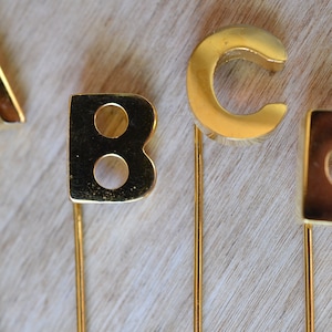 Peut inclure: Quatre épingles en forme de lettres dorées avec les lettres A, B, C et D. Les épingles sont sur une surface en bois clair.