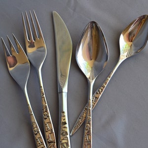 May include: A set of stainless steel silverware including two forks, a knife, and two spoons. The handles have a textured, geometric design. The silverware is arranged on a gray surface.