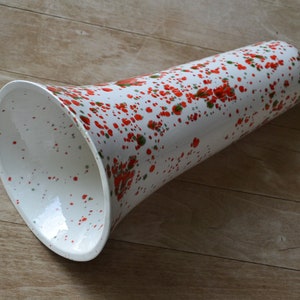 May include: White ceramic vase with a flared opening and a cylindrical body. The vase is decorated with red and green speckled paint. The vase is on a wooden surface.