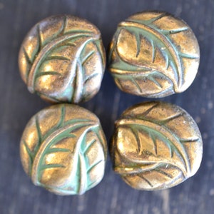 May include: Four round, gold-colored beads with a green leaf design. The beads are smooth and have a slightly raised design.