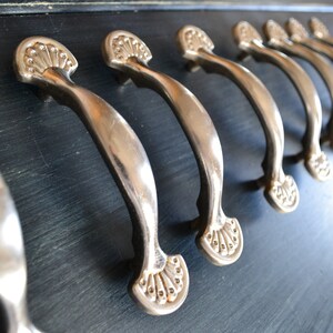May include: A series of metallic silver drawer pulls with a curved handle and a fan-shaped decorative base at each end. The hardware is mounted in a row on a dark, wood-grained surface, showing a consistent design across all the individual pieces.
