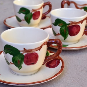 May include: Set of four white teacups with brown handles and a brown, green, and red apple design. The teacups are on matching saucers.