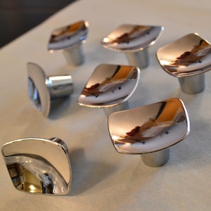 May include: Seven shiny silver cabinet knobs with a square shape and rounded edges. The knobs have a reflective surface and are arranged on a white surface.