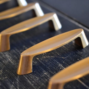 May include: Five gold-toned cabinet pulls with an oval shape and a slightly curved design. The pulls are arranged in a row on a dark wood surface.