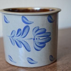 May include: A white ceramic crock with a brown rim and blue floral design. The design features a stylized flower with leaves and scattered petals.