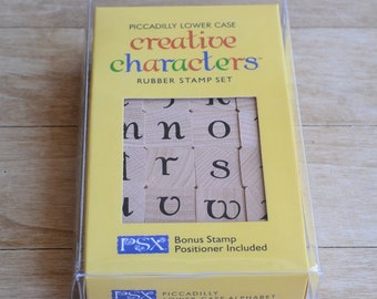 May include: A yellow package containing a set of lowercase alphabet rubber stamps. The stamps are arranged in a grid and feature a variety of creative fonts. The package includes a bonus stamp and a positioner. The text on the package reads "Piccadilly Lower Case Creative Characters Rubber Stamp Set".