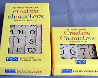 May include: Two yellow boxes with clear windows showing sets of rubber stamps. The top box contains lowercase letters and the bottom box contains numbers. Both boxes have the text "Creative Characters" and "Rubber Stamp Set" on them.